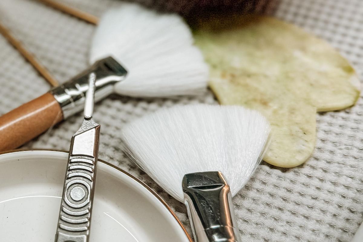 A dermaplaning blade, two fan brushes, and a gua sha stone laid out before a treatment