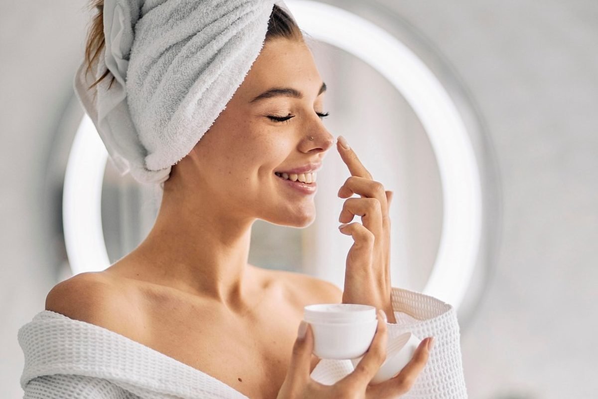 A woman in a robe and hair towel dabbing cream onto her nose from a small white jar, a lit round mirror behind her