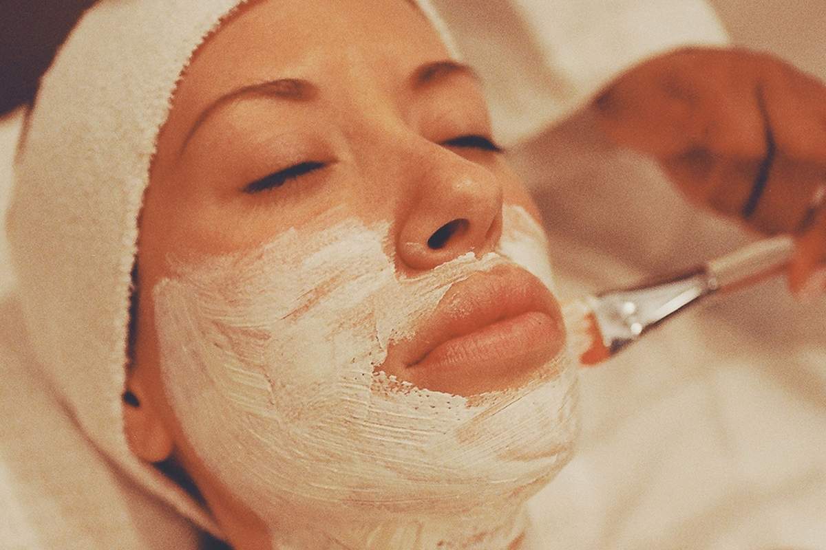 A woman resting with her eyes closed, a treatment mask brushed across her cheek, jaw and neck, her hair wrapped in a towel
