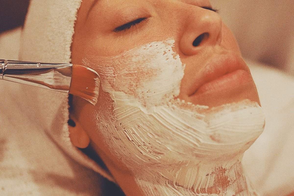 A brush laying a white treatment product across a client's cheek, jaw and neck, her eyes closed