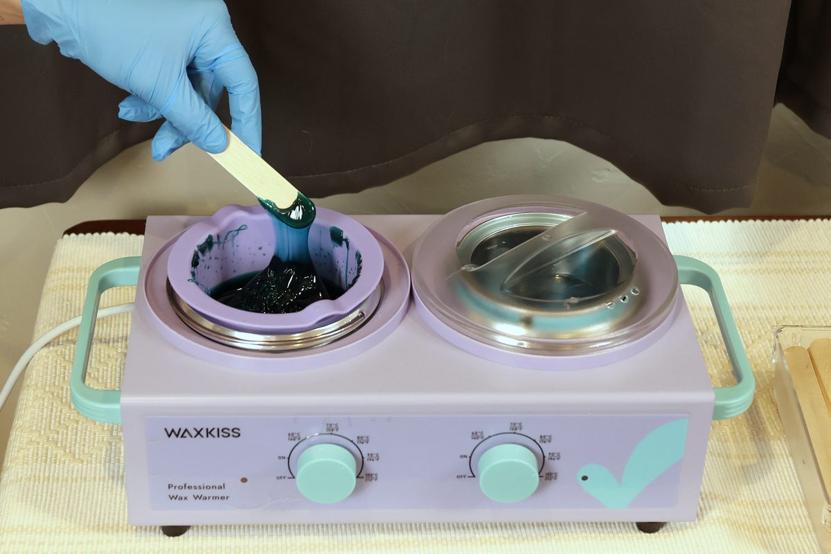A gloved hand lifting warm wax from the warmer on a spatula